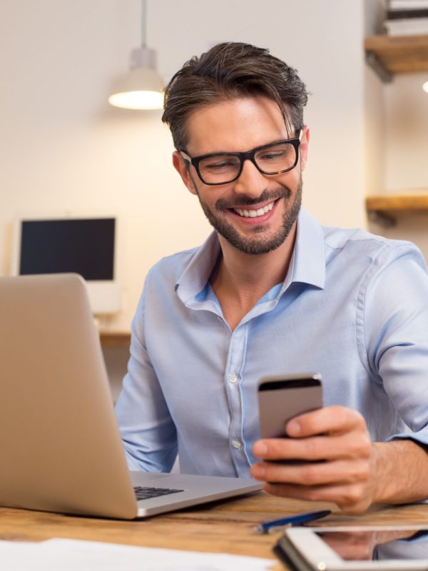 Young happy businessman smiling while reading his smartphone. Portrait of smiling business man reading message with smartphone in office. Man working at his desk at office.